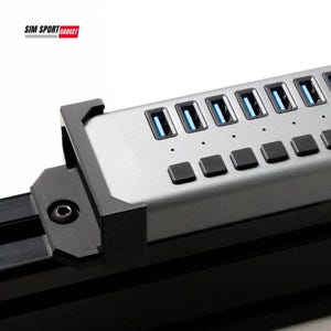 Puede incluir: Un concentrador USB plateado con múltiples puertos y botones negros. El concentrador tiene puertos USB iluminados en azul y está montado en un soporte negro. El logotipo "SIM SPORT GADGET" es visible en la esquina superior izquierda.