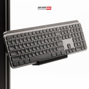 Puede incluir: Un teclado inalámbrico gris con teclas negras y una base negra. El teclado tiene un diseño completo con teclas numéricas, teclas de función y teclas de flecha. La marca "Logi" es visible en la parte superior. El texto "SIM SPORT GADGET" está en la esquina superior derecha.