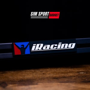 Puede incluir: Un logotipo iRacing en blanco y negro con un icono de piloto de carreras rojo, blanco y azul sobre un fondo negro. El texto "SIM SPORT GADGET" está por encima del logotipo.