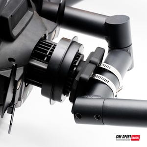 May include: Close-up of a black mechanical device with a cylindrical component and a fan-like structure. The device is attached to a black metal frame with metal clamps. The image is set against a white background.