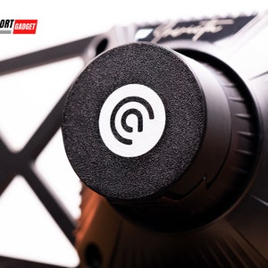 May include: Close-up of a black circular button with a white logo featuring a stylized "@" symbol. The button is textured and has a slightly raised edge.
