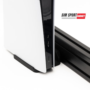 May include: A white PlayStation 5 console with a black side panel is mounted on a black metal stand. The console is sitting on a white surface. The stand has the text "SIM SPORT GADGET" printed on it.