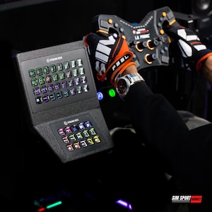 Puede incluir: Un Stream Deck negro con múltiples botones iluminados, colocado cerca de un volante de carreras. El Stream Deck tiene dos secciones, cada una con una cuadrícula de botones coloridos y personalizables. El volante es negro y naranja.