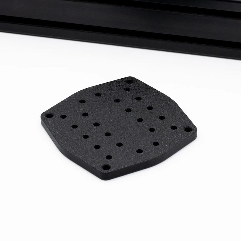 May include: A black, diamond-shaped metal plate with multiple evenly spaced holes. The plate has a textured surface and rounded edges. A black, rectangular object is visible in the background.