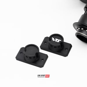 May include: Two black mounting brackets with a circular top and rectangular base. One bracket has the white text "VTS" on the top. The image also shows a portion of a black device with silver accents. The text "SIM SPORT GADGET" is at the bottom.