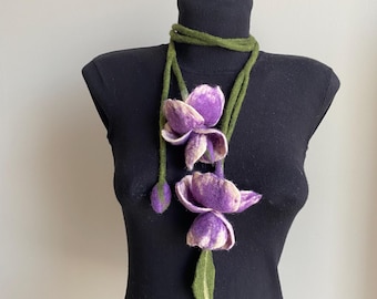 May include: A handcrafted necklace featuring felted purple tulip flowers with white accents and green stems and leaves. The necklace is displayed on a black mannequin, showcasing the floral design's intricate details and color palette.