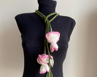 May include: A felted floral necklace featuring three white and pink flower buds with green stems and leaves. The necklace is displayed on a black turtleneck top.