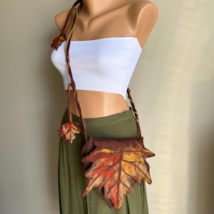 Könnte beinhalten: Eine braune Filz-Umhängetasche in Form eines Herbstblattes mit roten, orangefarbenen und gelben Farbtönen. Die Tasche hat einen langen, gedrehten Riemen und ein kleines Blattdetail. Die Tasche wird auf einer Schaufensterpuppe präsentiert.