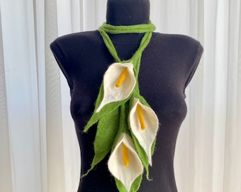 Può includere: Una collana di fiori in feltro verde e bianco con tre calle bianche e foglie verdi. La collana è esposta su un manichino nero.