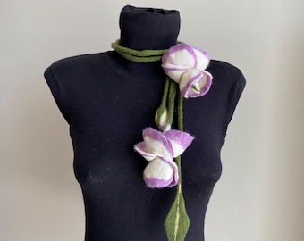 May include: A felted floral necklace displayed on a black mannequin. The necklace features white and purple flower buds, a green stem, and a green leaf. The mannequin is on a wooden stand.