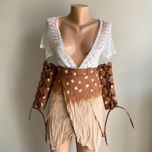 May include: A white lace top with a deep V-neck is paired with a brown and white spotted wrap skirt and matching arm cuffs. The skirt is layered with a beige fabric, creating a unique, bohemian-inspired outfit.
