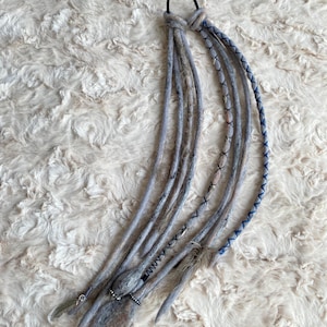 May include: Gray and blue dreadlock extensions with a black hair tie. The dreadlocks have decorative stitching in black and blue, with silver-colored beads and a feather charm. The extensions are made of a felted material.