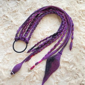 May include: Braided purple and black hair extensions with a black elastic. The extensions have pink accents, beaded details, and a decorative charm. The hair accessory is set against a fluffy, off-white background.