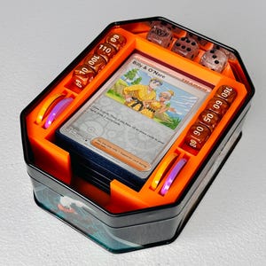 May include: A black metal storage tin containing a deck of trading cards, including a Billy & O'Nare trainer card, alongside orange damage counter dice marked with numbers, translucent purple and orange circular tokens, and clear dice.