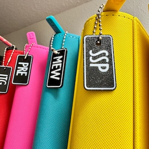 May include: Four colorful zippered pouches with black and white labels hanging from a shelf. The labels read "TIC", "PRE", "MEW", and "SSP".