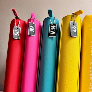 May include: Five colorful binders with metal zipper pulls and small metal tags with the words "JTG", "PRE", "MEW", "SSP" on them.