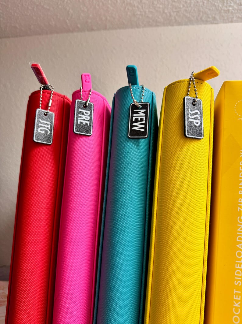 May include: Five colorful binders with metal zipper pulls and small metal tags with the words "JTG", "PRE", "MEW", "SSP" on them.