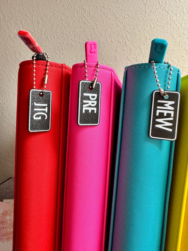 May include: Four colorful binders with zipper pulls and metal keychains with tags that read "JTG", "PRE", "MEW", and an unreadable word. The binders are red, pink, blue, and yellow.