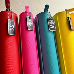 May include: Four colorful zippered binders with black metal tags hanging from them. The tags have the words "JTG", "PRE", "MEW", and "SP" written on them.