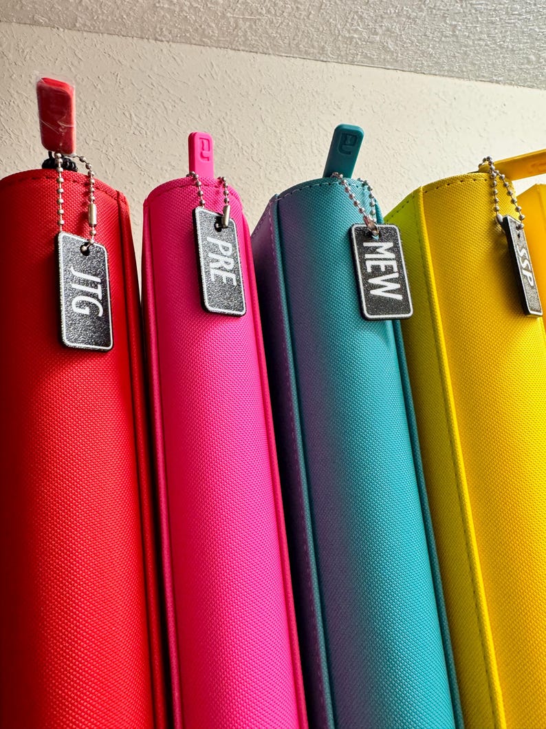 May include: Four colorful zippered binders with black metal tags hanging from them. The tags have the words "JTG", "PRE", "MEW", and "SP" written on them.