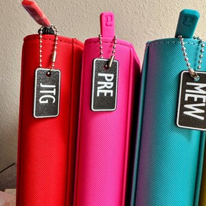 May include: Three zippered pouches in red, pink, and teal with black and white labels that say "JTG", "PRE", and "MEW" respectively.