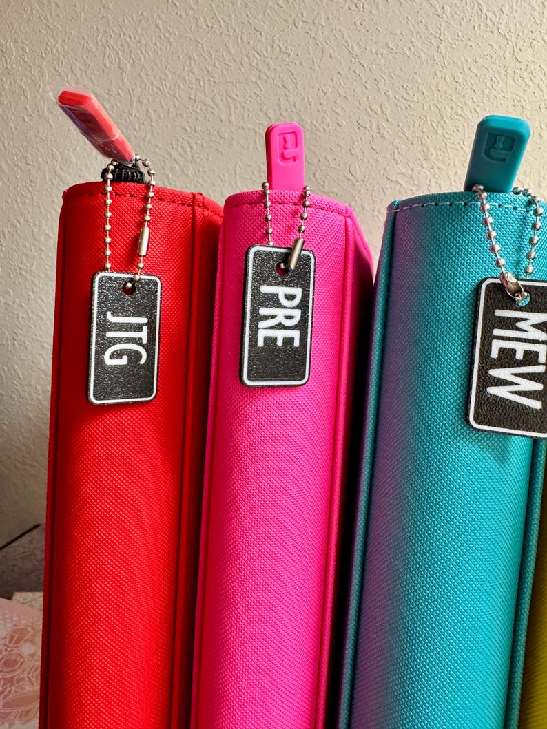 May include: Three zippered pouches in red, pink, and teal with black and white labels that say "JTG", "PRE", and "MEW" respectively.