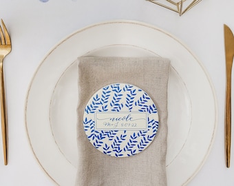 May include: A white plate with a blue and white patterned coaster with the name "nicole" and the date "May 8, 2022" written on it. The coaster is on a beige linen napkin. There is a gold fork and knife on the white table cloth. There are two gold geometric candle holders with white candles.