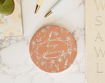 Könnte beinhalten: Ein runder Untersetzer aus Terrakotta mit einem weißen Blumenmuster und dem Text "keep growing" in weißer Schrift. Der Untersetzer liegt auf einer weißen Marmorfläche mit zwei Stiften und einem Notizblock, auf dem in weißer Schrift "WRITE SLOW AND WITH LOVE" steht.