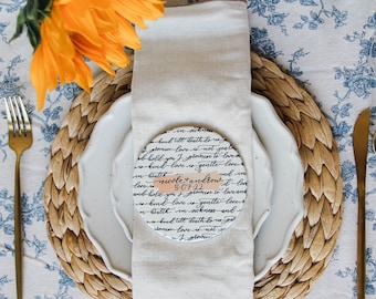 May include: A white ceramic plate with a black and white handwritten design on a round coaster. The plate has a handwritten message that says "love is love is love is love" and "Nicole + Andrew 8.07.22". The plate is set on a woven straw coaster. Two gold forks and two gold spoons are on a white tablecloth with a blue floral pattern.