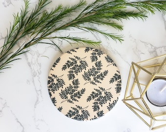 May include: A round coaster with a black and cream floral pattern, a gold geometric candle holder with a white tea light candle, and a sprig of green foliage on a white marble surface.