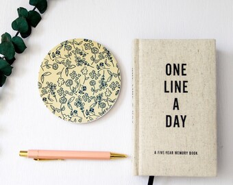 May include: A beige journal with the text "One Line A Day" and "A Five-Year Memory Book" on the cover. The journal is sitting on a white surface next to a coaster with a floral pattern.