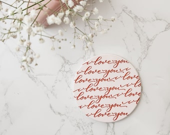 Könnte beinhalten: Ein runder weißer Untersetzer mit dem Satz "i love you" in roter Kursivschrift.