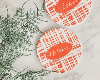 Könnte beinhalten: Zwei runde Untersetzer mit einem weiß-orangen Karomuster. Jeder Untersetzer hat ein oranges Oval mit einem Namen in Schreibschrift: "Parker" und "Norrin". Die Untersetzer liegen auf einer weißen Marmoroberfläche mit grünen Zweigen.