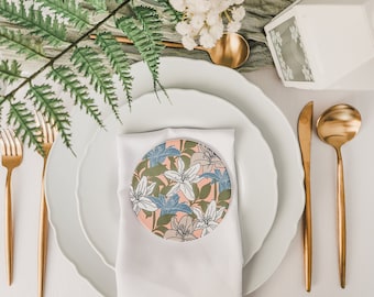 May include: A white napkin with a floral design coaster on top of it. The coaster is round and has a pink background with blue and green flowers. The napkin is on top of two white plates. There are gold forks and a gold spoon on the table.