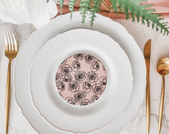 May include: A white dinner plate with a pink floral design coaster in the center. The plate is set on a white marble surface with gold flatware and a green fern.