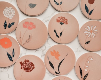 Könnte beinhalten: Set mit 11 runden Untersetzern mit Blumenmotiven. Jeder Untersetzer zeigt eine andere Blume in Rot-, Orange-, Weiß- und Brauntönen auf einem hellrosa Hintergrund.