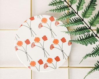 Könnte beinhalten: Ein runder weißer Untersetzer mit einem sich wiederholenden Muster aus orangefarbenen Blumen auf weißem Hintergrund. Der Untersetzer befindet sich auf einer weißen Oberfläche mit einem goldenen Gitter.