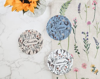 Könnte beinhalten: Drei runde Untersetzer mit Blumenmuster und den Namen "Kristina", "Samantha" und "Cassandra" in Kursivschrift. Die Untersetzer befinden sich auf einer weißen Marmorfläche mit einer Sonnenblume in einer Glasvase und einer Leinentasche mit gestickten Blumendesigns.