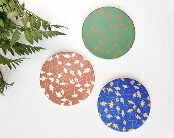 Könnte beinhalten: Drei runde Untersetzer mit einem Blumenmuster. Die Untersetzer sind grün, braun und blau mit goldenen Blumenmustern.