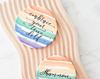 May include: Two round coasters with a rainbow watercolor design. The top coaster says "embrace your true self" in black calligraphy. The bottom coaster says "Shannon" in black calligraphy.
