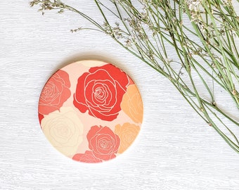 May include: A round coaster with a floral design featuring red, orange, and white roses on a pink background.