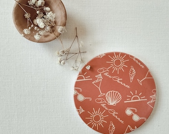 Könnte beinhalten: Ein runder Untersetzer in Terrakottafarbe mit einem weißen Design aus Sonnen, Sonnenbrillen, Eisbechern, Muscheln und dem Text "Ich liebe den Sommer".