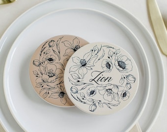 May include: Two round, floral-patterned coasters with black ink on a beige background. One coaster has the name "Zion" written on it.