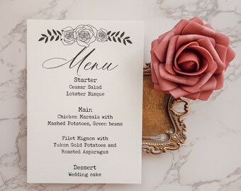 May include: A white menu card with a floral design and the word "Menu" written in cursive. The menu lists the following items: Starter - Ceasar Salad, Lobster Bisque; Main - Chicken Marsala with Mashed Potatoes, Green beans, Filet Mignon with Yukon Gold Potatoes and Roasted Asparagus; Dessert - Wedding cake.