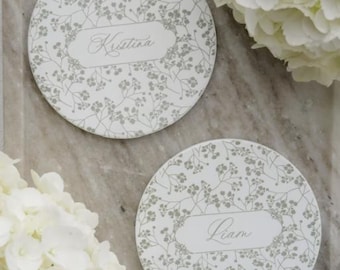 Könnte beinhalten: Zwei runde Untersetzer mit floralem Muster und den Namen "Kristina" und "Liam" in Schreibschrift. Die Untersetzer sind weiß mit einem grauen Blumenmuster, neben weißen Hortensien.