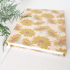 May include: A pink and white journal with a sunflower pattern. The cover is decorated with a repeating pattern of yellow sunflowers with brown centers on a light pink background.