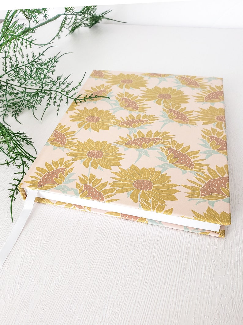 May include: A pink and white journal with a sunflower pattern. The cover is decorated with a repeating pattern of yellow sunflowers with brown centers on a light pink background.