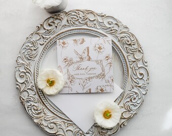 May include: A white and gold floral "Thank you" card with a butterfly design, placed on a decorative, ornate, white-washed, round tray. The tray is surrounded by white flowers. The card reads "Thank you with love, Vanessa."