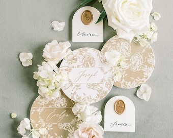May include: Elegant wedding place cards and floral arrangements. The design features white and gold floral patterns, with names like "Elvira", "Joseph", "Evelyn", and "James" written in script. White roses and hydrangeas are arranged on a soft green background.