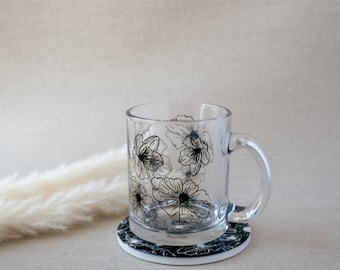 Könnte beinhalten: Eine klare Glas-Tasse mit einem schwarzen Blumenmuster steht auf einem schwarzen und weißen Blumen-Untersetzer. Die Tasse hat einen Henkel und ist leer.